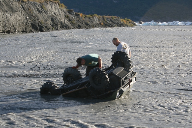 ATV overturned in shallow river with two people nearby
