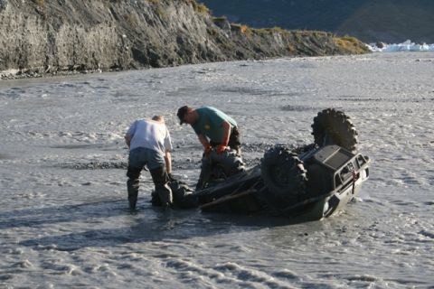 1990 Chevrolet Suburban - An ATV has overturned in a shallow, muddy river, with two individuals attempting to recover it.