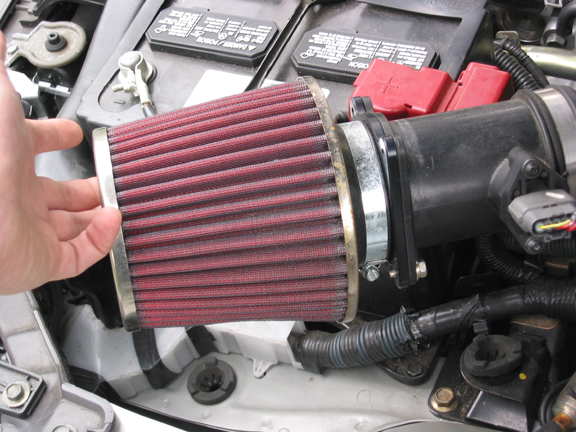 Hand holding a red cone air filter clamped to a black intake tube in an engine bay