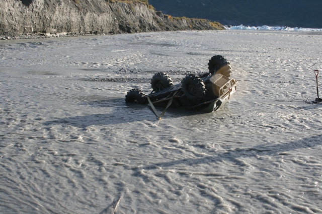 ATV flipped on its side in a shallow, muddy river with a tow strap attached. A shovel stands in the background