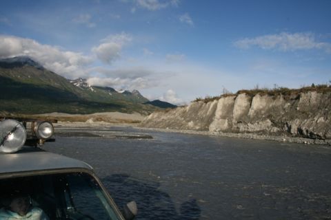 1990 Chevrolet Suburban - A vehicle with auxiliary lights is positioned in a rocky riverbed, with a dramatic cliff face and mountains under a cloudy sky.