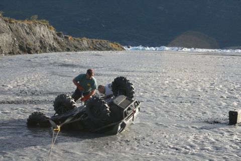 1990 Chevrolet Suburban - Two individuals work to recover an overturned ATV from a shallow, rocky riverbed with a mountainous backdrop.