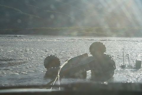 1990 Chevrolet Suburban - A vehicle is stuck in a river, partially submerged and overturned, with shovels visible in the background.