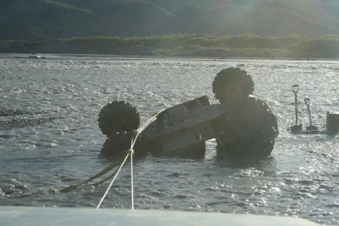 1990 Chevrolet Suburban - A vehicle is stuck in a river, partially submerged and tilted to its side, with a tow rope attached.