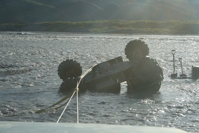 Tilted vehicle submerged in river, with large tires visible and a tow rope attached