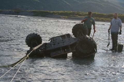 1990 Chevrolet Suburban - Two men attempt to recover an overturned ATV from a shallow riverbed, with a tow strap attached.