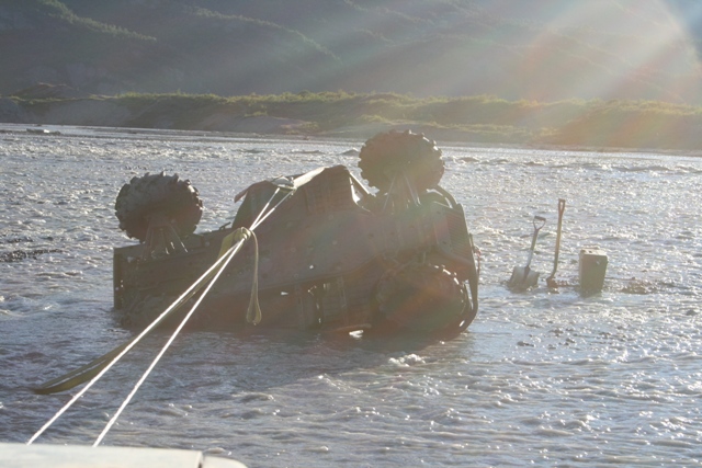 Overturned vehicle with large tires being pulled from shallow water by ropes; shovels and a bucket visible on shore