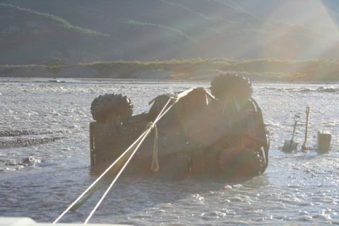 1990 Chevrolet Suburban - An overturned vehicle is stuck in shallow water, with recovery equipment like shovels visible on the bank.