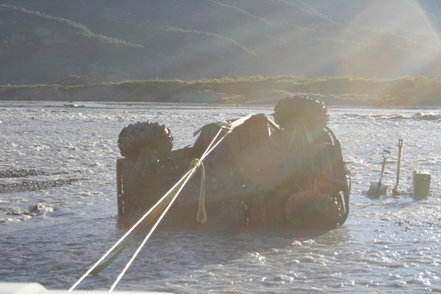 vehicle flipped on its side in shallow water, being pulled by ropes. Shovels and a toolbox are nearby