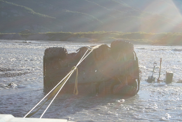 Overturned dark-colored vehicle in shallow water with tow ropes attached; two shovels and a block visible on shore