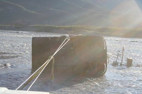 1990 Chevrolet Suburban - An overturned vehicle is submerged in shallow water, with straps attached for recovery. Shovels lie in the water nearby.
