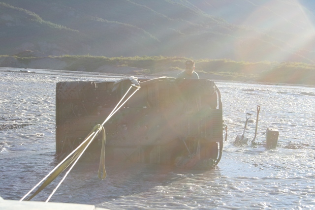 Vehicle partially submerged in a river, being pulled by ropes. Shovels are stuck in the riverbed nearby
