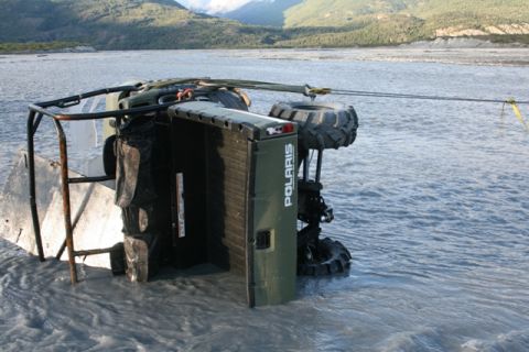 1990 Chevrolet Suburban - An olive green Polaris ATV is overturned and partially submerged in a river, with a tow rope attached.