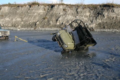 1990 Chevrolet Suburban - A white pickup truck tows a green ATV out of a shallow, muddy river with a steep bank in the background.
