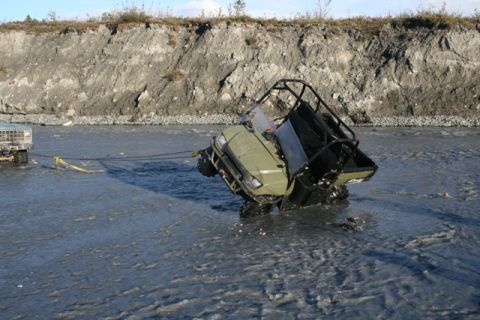 1990 Chevrolet Suburban - A green ATV is being towed out of a shallow river by a truck, with a steep embankment in the background.