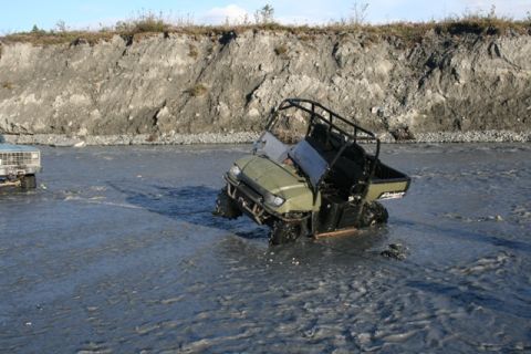 1990 Chevrolet Suburban - Two vehicles navigate a shallow, muddy riverbed with a steep embankment in the background.