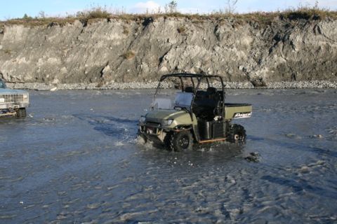1990 Chevrolet Suburban - Two vehicles navigate a shallow, rocky riverbed under a steep, eroded embankment.