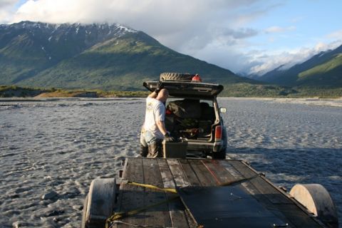 1990 Chevrolet Suburban - A person loads gear into an SUV parked in a shallow river, with a trailer attached and mountains in the background.