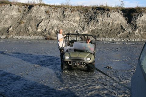 1990 Chevrolet Suburban - An ATV is being towed through a shallow river with a steep embankment in the background.