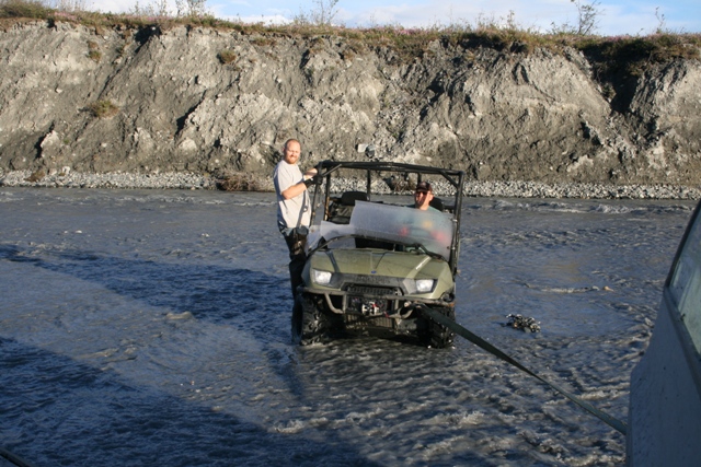 Guy standing next to an ATV stuck in a river, another vehicle is towing it out