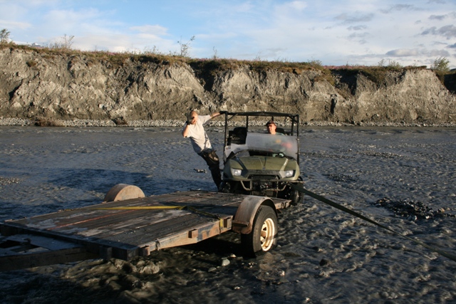 Green utility vehicle towing a trailer in a riverbed with two people