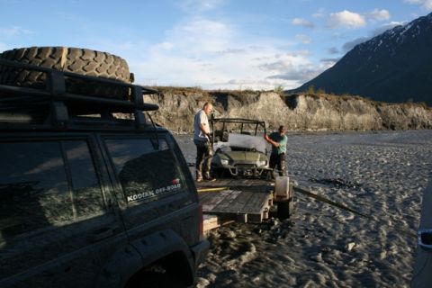 1990 Chevrolet Suburban - Two men load an off-road vehicle onto a trailer in a muddy, rocky area near a river.
