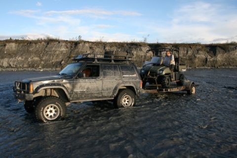 1990 Chevrolet Suburban - A Jeep Cherokee and a utility vehicle on a trailer navigate a rocky, shallow riverbed under a partly cloudy sky.