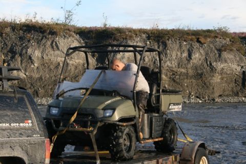 1990 Chevrolet Suburban - A man works on a utility vehicle's windshield while it is on a trailer, with a river and rocky bank in the background.