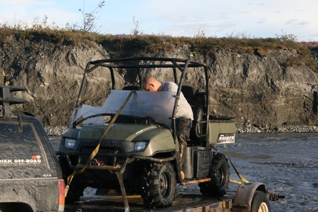 Green utility vehicle with a damaged windshield on a trailer next to a black SUV
