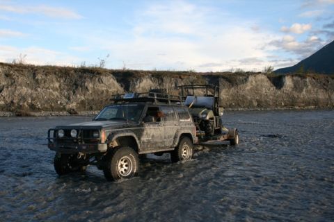 1990 Chevrolet Suburban - A modified Jeep Cherokee navigates a rocky riverbed, pulling a trailer carrying an ATV.