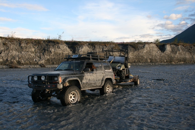 Off-road Jeep Cherokee towing a trailer with an ATV through a shallow river