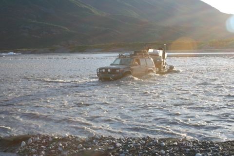 1990 Chevrolet Suburban - A Jeep Cherokee XJ navigates a river while towing a trailer, with a mountainous landscape in the background.