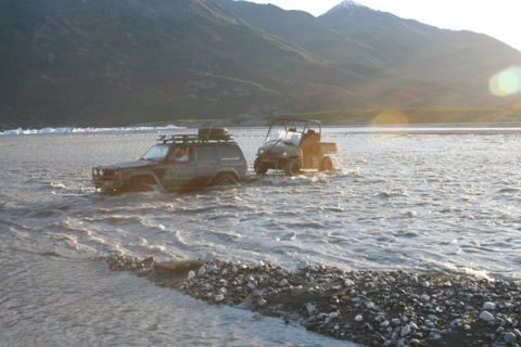 1990 Chevrolet Suburban - Two off-road vehicles navigate a wide, shallow riverbed under a bright, setting sun.