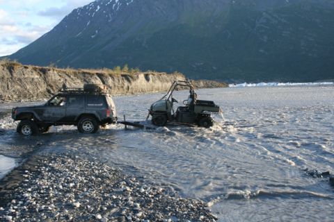 1990 Chevrolet Suburban - Two off-road vehicles navigate a shallow river with a mountain range in the distance.