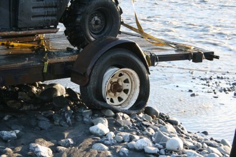 1990 Chevrolet Suburban - A trailer sits on a rocky shore with a flat tire, partially submerged in shallow water.