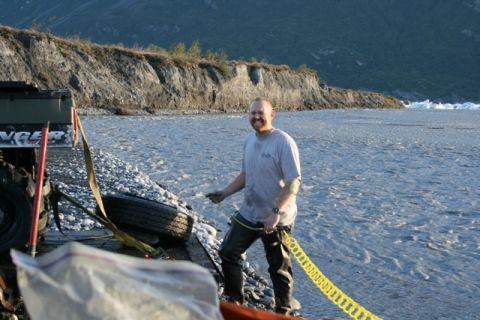 1990 Chevrolet Suburban - A man stands on a rocky shore, holding a yellow recovery strap near a vehicle.