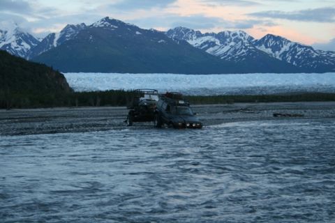 1990 Chevrolet Suburban - A dark-colored SUV with a trailer is driving through a shallow river with a large glacier and snow-capped mountains in the background.