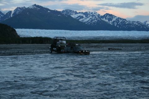 1990 Chevrolet Suburban - A dark-colored SUV is fording a shallow river with an ATV trailer attached, with a vast glacier and snow-capped mountains in the background.