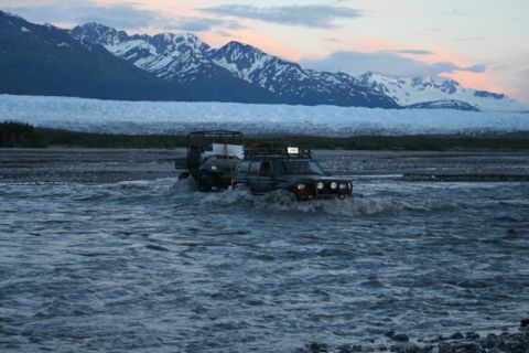 1990 Chevrolet Suburban - Two off-road vehicles, a dark SUV and a smaller ATV, drive through a wide, rocky river with a large glacier and snow-capped mountains in th…