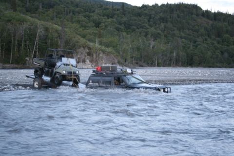 1990 Chevrolet Suburban - A black SUV is partially submerged in a rocky river, being towed by a green utility vehicle. The SUV has a roof rack loaded with gear and i…