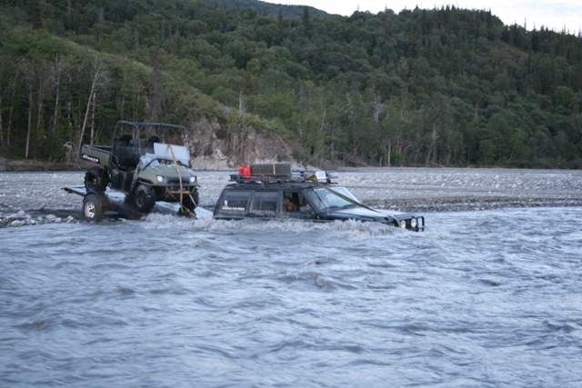 Black SUV submerged in a river, being towed by a green utility vehicle.