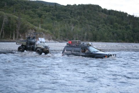 1990 Chevrolet Suburban - Two off-road vehicles are fording a shallow, fast-moving river. A black SUV with a roof rack is being towed by a green utility vehicle.