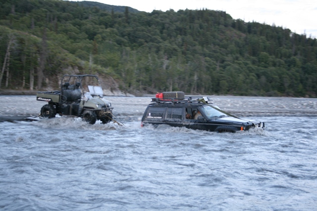 Black SUV with roof rack being towed by green utility vehicle through a river.