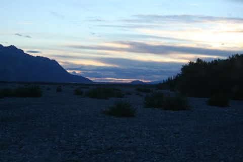 1990 Chevrolet Suburban - A wide shot of a rocky riverbed with sparse vegetation under a twilight sky filled with colorful clouds. A large mountain range is visible…