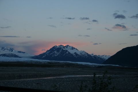 1990 Chevrolet Suburban - A mountain range is visible at dusk, with the sun setting behind the central peak, casting a pink glow on the clouds. A wide, rocky riverbe…