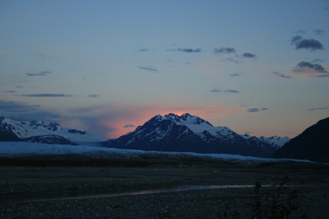 Mountain range at sunset with snow-capped peaks and a rocky foreground.