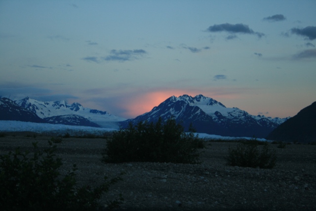 Mountain range at sunset with snow-capped peaks and a rocky foreground.