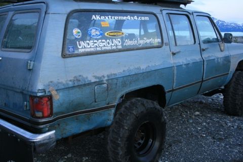 1990 Chevrolet Suburban - The rear passenger side of a light blue, lifted Chevrolet Suburban is shown parked on a gravelly surface with mountains in the background.…