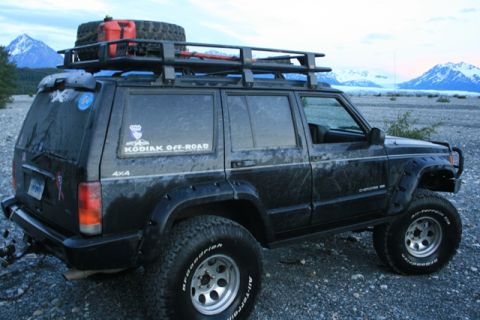 1990 Chevrolet Suburban - A black Jeep Cherokee is parked on a gravelly, rocky terrain with snow-capped mountains in the background. The vehicle has a roof rack with…