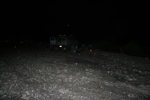 1990 Chevrolet Suburban - A group of people are gathered around a campfire at night next to a dark-colored SUV with its doors open. The vehicle is parked on a rocky,…